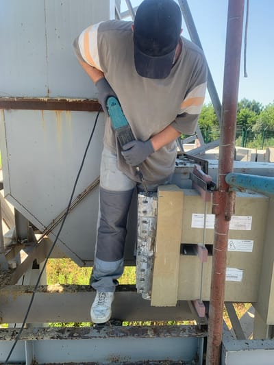 A person, wearing a grey t-shirt with orange and white stripes on the sleeves, dark pants with grey panels, grey work gloves, a black baseball cap, and white sneakers, is using a blue reciprocating saw on a light grey metal structure. The person is positioned on a metal platform, with a large, light grey industrial structure to their left. To the right of the person, a light brown rectangular object with labels is visible, supported by metal beams. A rusty red pipe stands vertically in front of this object. The background shows green trees and a clear blue sky. This scene is located at the South Ukrainian Nuclear Power Plant in Pivdennoukrainsk, Ukraine, likely during maintenance or construction work.