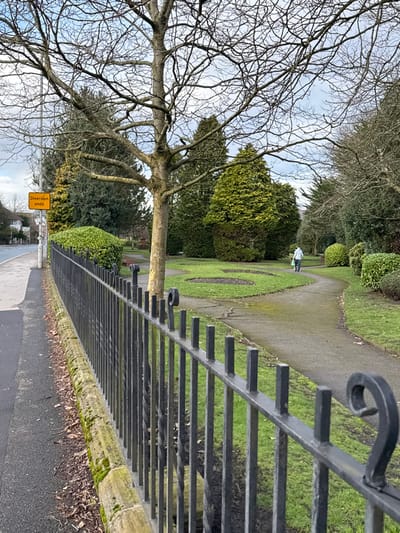 The image depicts a public green space adjacent to a paved road, located within the district of West Lancashire, United Kingdom.

In the foreground, a black wrought iron fence with decorative scrolls at intervals extends diagonally from the lower left towards the mid-right. A moss-covered stone kerb runs along its base. To the left of the fence is a dark asphalt pavement, beyond which lies a two-lane asphalt road. A yellow rectangular "Diversion ends" sign on a pole is visible on the roadside, left of the central tree. In the far left background, residential buildings are partially visible across the road.

The green space features well-maintained grass areas and several winding dark grey asphalt paths. Two distinct circular patches of bare earth, suggestive of flowerbeds, are present in the grassy areas. A prominent deciduous tree with bare branches stands centrally. Numerous evergreen conifer trees, including tall conical specimens and rounded shrubs, are scattered throughout the park, alongside other bare-branched deciduous trees, indicating a late autumn or winter season.

One individual is observed walking away from the viewer on a winding path within the park, heading towards the right side of the frame. They are wearing light-colored clothing on their upper body and dark trousers, carrying a light green object in their left hand. The sky is partially cloudy and bright.