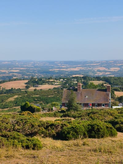 The image depicts a panoramic view from an elevated position in Cleehill, United Kingdom, showcasing a rural landscape under a clear blue sky. In the mid-ground, a prominent brick house with a tiled roof and two chimneys stands on a hillside covered in dense green vegetation. The house has several windows, and a red sign is visible in one of them. The surrounding terrain is characterized by rolling hills with patches of dry grass, fields of various shades of brown and green, and clusters of trees. In the background, a vast expanse of countryside stretches to the horizon, featuring more undulating hills and agricultural fields. The overall scene conveys a sense of tranquility and rural serenity. There are no people or animals visible in the image.