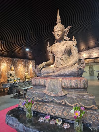 In the city of Na Chom Thian, Thailand, an indoor scene features a large, ornate statue of Buddha seated in a meditative pose. The Buddha statue is made of metal, with intricate carvings and colorful embellishments. In the foreground, a vase of lotus flowers and a few candle holders are placed on a dark marble surface. Behind the main Buddha, smaller Buddha statues are visible, as well as a mosaic wall depicting figures. The ceiling of the room is made of dark wood slats. The overall atmosphere suggests a temple or a museum dedicated to Buddhist art.