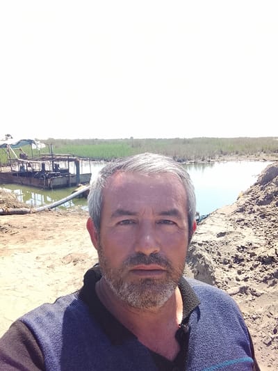 A man is taking a selfie. He has gray hair and a beard. He's wearing a blue sweater. In the background, there's a body of water with reeds growing on the far side. There is a crude metal structure of some kind on the water, possibly a small dredge or pump platform. The ground is sandy and muddy. It is daytime and a slightly overcast day.
It is not possible to confirm the location is in Syrdarya, Uzbekistan, based on the visual information provided in the image.