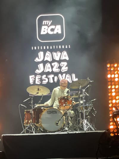 The image captures a performance at the International Java Jazz Festival, likely in Salembaran, Indonesia. The backdrop displays the festival's logo featuring "my BCA" and "JAVA JAZZ FESTIVAL INTERNATIONAL" in large, white lettering against a black background. A drummer is positioned behind a complex drum set with multiple cymbals and toms. The drum set includes a bass drum with "wave Earth" logo on its head. The lighting above the stage consists of numerous circular lights. The overall environment suggests a concert setting.