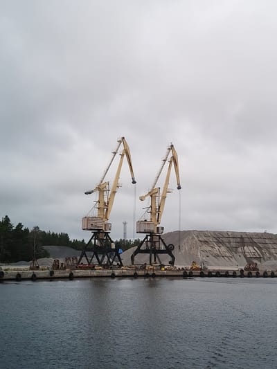 The image depicts a port scene in Shal'skii, Russia, under an overcast sky. Dominating the mid-ground are two large, yellow gantry cranes positioned on a concrete pier. The cranes have intricate metal structures and long, angled jibs. To the right of the cranes, a massive pile of sand or gravel is visible, with terraced layers indicating recent material handling. In the foreground, dark, rippling water stretches across the frame, with a section of the pier visible along the bottom edge. The pier is lined with dark, circular buoys and a few pieces of industrial equipment, including what appear to be smaller cranes or loading machines. A line of dark green trees borders the background on the left, beyond the pier and material piles. A tall, slender tower, possibly a communication tower, is visible in the distance behind the cranes. The overall impression is one of an industrial port engaged in bulk material handling.
