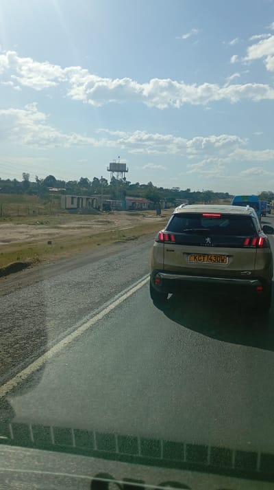 A tan Peugeot SUV occupies the right lane of a highway. The license plate reads "KCT 430W". To the left of the road, a sparse landscape stretches towards a cluster of small buildings and a tall water tower. The sky is pale blue, dotted with puffy white clouds and a faint glare from the sun. The viewer is likely inside a vehicle, as evidenced by the partial view of a dashboard at the bottom of the frame.