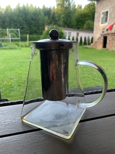 A glass teapot with a black lid and infuser sits on a dark brown, slatted surface. The teapot has a square base and a curved glass handle. Water droplets are visible on the exterior of the teapot. In the background, a well-maintained green lawn stretches out, bordered by a line of trees. Further back, a light-colored building with a white-framed window and a red cloth hanging on its exterior is partially visible, suggesting a residential area. The scene appears to be set outdoors during daylight hours, possibly in a garden or patio area. The location is identified as Silmala, Latvia.