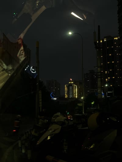 Captured at night in 龙溪街道, China, the image shows a cityscape illuminated by artificial lights. Tall buildings with multiple lit windows dominate the backdrop. A street lamp casts a bright glow on the scene. The foreground features objects suggesting a roadside perspective, including a partially visible vehicle and other urban elements.