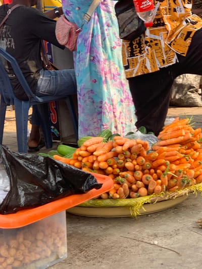 The scene depicts a street market setting in Ebute-Metta, Nigeria. In the foreground, a large, shallow, circular woven tray contains numerous bright orange carrots, many appearing bundled. To the left and behind the carrots, several green cucumbers are visible. Further left, a clear plastic container with an orange lid is partially visible, holding light brown granular items, possibly nuts or snacks, with a crumpled black plastic bag resting on its lid. The ground is a light gray concrete surface.
Three individuals are partially visible. On the left, a person is seated on a blue plastic chair, wearing a dark long-sleeved shirt with a light-colored graphic on the back and blue denim jeans. A brown strap crosses their torso. In the center, a person stands, wearing a light blue dress or wrapper with purple and pink floral patterns. A light-colored strap extends over their shoulder, and a pink woven purse hangs at their side. On the right, another person stands, wearing a yellow and orange patterned short-sleeved shirt over dark trousers. A red Coca-Cola bottle is visible near their waist, and a dark fanny pack or small bag is present on their right hip. No direct interactions between individuals are depicted.