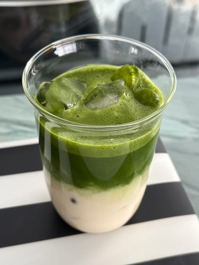 A close-up, high-angle shot depicts a clear glass of iced matcha latte. The matcha is layered over a creamy liquid at the bottom. Ice cubes are visible in the matcha layer. The glass sits on a black-and-white striped surface. The background appears to be a blurred light-colored interior space. The location is Zapopan, Mexico.