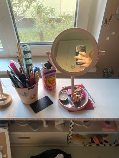 The image depicts a close-up, eye-level, indoor scene on a white wooden desk. The desk surface is cluttered with various personal items, suggesting a vanity or crafting area. Prominently featured is a pink, round mirror on a stand, reflecting a hand holding a smartphone. To the left of the mirror is a light pink bucket filled with pens, pencils, and other writing utensils. A pink can with a "Chupa Chups" label stands next to the bucket. In front of the bucket, a black card is laid flat. The mirror's tray contains a white candle, small toys including a cow and a pink character, and a small waffle-shaped object. Scattered on the desk are blue beads and red paper. The background shows a window with a view of greenery, suggesting a residential setting in Мухавецкі сельскі Савет, Belarus. Below the desk, drawers and shelves are visible, containing various items, including what appears to be a pink camera and cow-print patterned fabric. The overall lighting is natural, coming from the window, creating soft shadows and highlights.