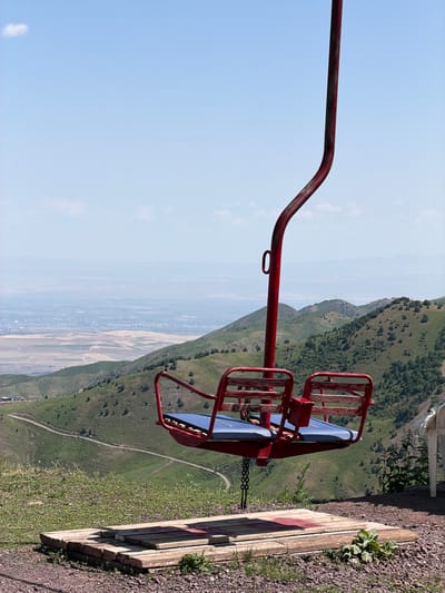 A red ski lift chair with two blue seats is suspended in the air against a clear blue sky. The chair hangs from a red metal pole that curves upwards. In the background, rolling green hills descend towards a city in the distance, with a hazy, muted cityscape visible under a pale sky. A winding dirt path is visible on one of the hills. In the foreground, a wooden platform made of several planks lies on the ground, surrounded by gravel and sparse green vegetation. A white chair is partially visible on the right edge of the frame. The scene appears to be a scenic overlook or a stop on a ski lift, likely in a mountainous region like Kashka Suu, Kyrgyzstan.
