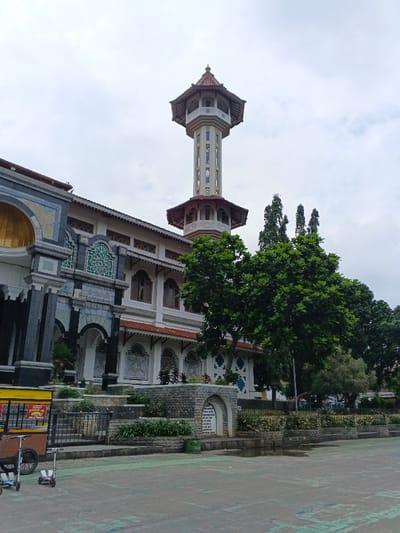 This image depicts a large architectural complex identified as a mosque, located in Bien Hoa, Vietnam, under a uniformly overcast sky. A prominent, tall, octagonal minaret with multiple open-air balconies and a tiered, reddish-brown tiled roof rises centrally. The main building features white and light-colored facades, a multi-tiered reddish-brown tiled roof, and numerous arched windows with dark frames. Sections of the exterior are adorned with distinctive teal and white geometric patterns. A significant entrance on the left side of the building includes dark pillars and a golden-colored arched alcove. The foreground presents a paved plaza composed of grey and light green rectangular sections. On the far left, a brown wooden vendor cart with a yellow top and a red informational sign is parked near two child-sized kick scooters, one blue and one grey. A landscaped area separates the paved plaza from the building, featuring stone retaining walls, green shrubbery, a black metal fence, and a stone archway. A dark green cylindrical object is situated near the archway. Various trees with dense green foliage are distributed across the scene, some partially obscuring the building's lower levels and others rising behind its roofline. No individuals are present in the frame.