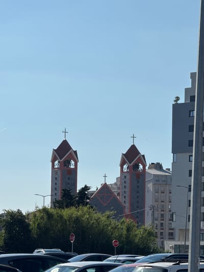 The image shows a clear, sunny day in a city, possibly Bjeliši, Montenegro. In the mid-ground, a church with two bell towers is visible. The towers are square with red, pointed roofs and crosses at the apex. The church also has a prominent red roof with a cross design. Behind the church, several modern buildings of varying heights and architectural styles are present. In the foreground, a row of cars is parked, and a stop sign is visible on the right side of the road. Lush green trees and bushes partially obscure the lower portion of the church and buildings. The sky is a clear blue with no clouds.