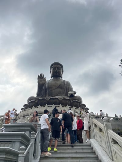 A large bronze Buddha statue dominates the view under a cloudy sky. A crowd of people is gathered on a staircase below the statue, some appearing to be taking pictures. The statue is viewed from a low angle, emphasizing its height. The architectural details of the staircase are visible in the foreground.