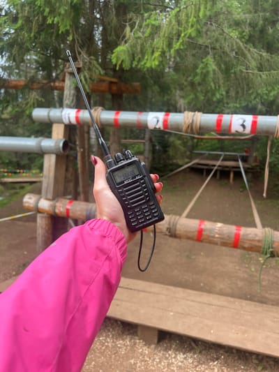 The media file shows a close-up of a person's hand holding a black walkie-talkie. The walkie-talkie is a VHF/UHF FM transceiver with a small display screen and several buttons. The screen displays "A-41" and some numbers and letters that are difficult to read. The buttons are labeled with functions like "SCAN," "MENU," "CALL," and "BEEP." The person's hand has dark red nail polish. They are wearing a bright pink jacket, the sleeve of which is visible on the right side of the frame.
The background suggests an outdoor setting, possibly a forest or park, with trees and wooden structures. There are vertical poles in the background, one of which is grey with red and white bands and a symbol that resembles a stylized heart or a number "3." Another pole is made of bamboo with red bands. The ground appears to be made of dirt and gravel. The lighting suggests it is daytime, and the overall atmosphere is casual, perhaps indicating an outdoor activity like hiking, a game, or work. The presence of the walkie-talkie implies communication is taking place or is about to take place. The location context provided is Сергино, Russia.