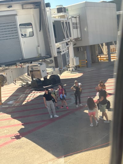 The image shows an airport scene. Six people are walking on the tarmac near a jet bridge connected to an aircraft. The jet bridge is white with the number "2400" displayed on the side. The tarmac has red lane markings. A multi-story building is visible adjacent to the tarmac. The location is likely Cinisi, Italy, based on external contextual information, though that is not apparent from the image itself.