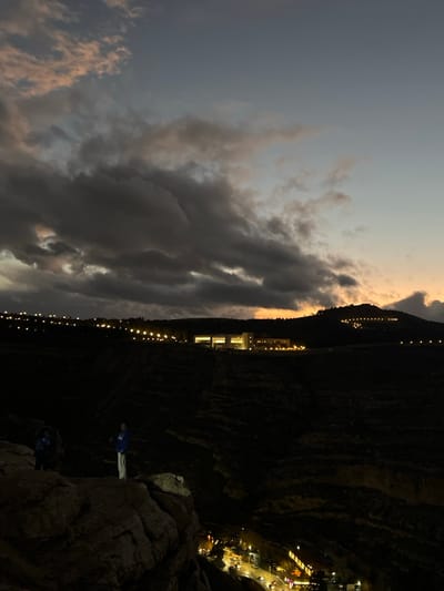 The image depicts a nighttime panoramic view of Dummar Municipality, Syria, at dusk. The sky is largely covered by dense, dark gray clouds, with a band of lighter blue-gray visible in the upper sky and a strip of warm orange and pink light from the setting sun along the eastern horizon.

In the immediate foreground, to the lower left, a large, rugged, brownish-gray rocky outcrop is present. Two individuals are standing on this outcrop. The person on the left is partially obscured, wearing a dark top. The second person, positioned more centrally, faces right, clad in a dark blue long-sleeved top and light-colored pants.

The midground consists of a steep, dark valley or ravine, largely obscured by shadow but revealing some textural details of rock and earth.

In the distant background, several illuminated features are visible across dark hills. A horizontal line of uniformly spaced, small yellow lights stretches across a hill on the left. Centrally, a prominent, multi-story building complex with numerous lit windows emits warm yellow light, positioned on a distant plateau. To the right of this complex, another hill ascends, marked by a curved line of smaller lights.

Below the valley in the lower right, a section of an urban area is visible, characterized by multiple points of artificial light from streetlights and vehicle headlights on a road, as well as illuminated windows of buildings.