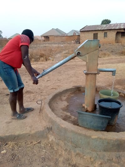 An outdoor scene depicts a person drawing water from a hand pump in a rural setting. The individual, seen from a side-rear angle, appears to be a man with dark skin, wearing a red short-sleeved t-shirt, blue denim shorts, and dark slide sandals. He is bent over, actively operating the long metal handle of the pump with both hands. His arms show muscular definition. He also has a dark cap or head covering.
The hand pump itself is made of metal, appearing sturdy and somewhat aged, possibly painted a light green or grey, with areas of discoloration or rust. Water is visibly flowing in a steady stream from its horizontal spout into a dark plastic bucket positioned within a circular concrete basin that surrounds the pump's base. Another lighter-colored bucket, already containing some water, and a wide, shallow dark plastic basin are also present in the concrete enclosure. The ground immediately around the pump and the person is dry, unpaved earth. A bent piece of metal, shaped like a hook, protrudes from the ground near the man's feet.
In the background, a cluster of simple, single-story houses with corrugated metal roofs can be seen. These structures appear to be built from local materials like mud brick. The surrounding landscape is characterized by dry, sparse yellowish vegetation and bare earth, suggesting an arid or semi-arid environment. The sky is a pale, hazy blue, indicative of daylight, possibly morning or late afternoon due to the soft lighting. The overall impression is that of a village scene where a hand pump serves as a communal water source. There is no visible text in the image.