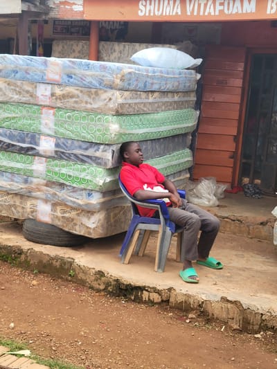 The image depicts an outdoor scene in Tangu, Nigeria, outside a shop. A young Black male is seated on a blue plastic chair, wearing a red t-shirt and grey pants, with green flip-flops. He is positioned in front of a tall stack of mattresses wrapped in clear plastic. The mattresses are of various patterns, including floral designs and a green pattern with circles. A white pillow rests on top of the mattress stack. The shop behind the mattresses has an orange awning with white lettering that appears to read "SHUMA VITAFOAM". Part of the shop's exterior is made of wooden planks. In the foreground, the ground is uneven, with dirt, small stones, and patches of grass. A tire is visible beneath the bottom mattress. The overall scene suggests a commercial setting, likely a mattress shop or distributor.