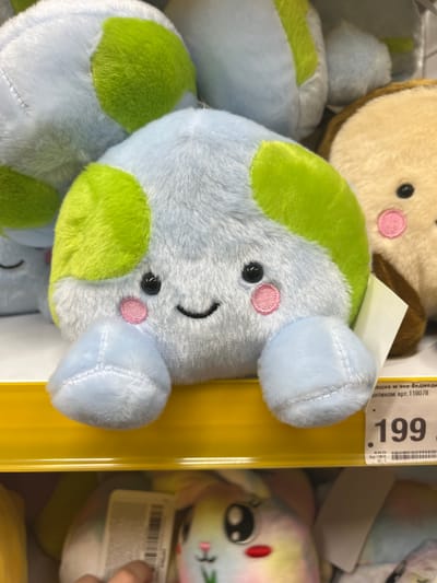 A close-up shot shows a row of plush toys on a yellow shelf. The main subject is a light blue, spherical plush toy with green patches and embroidered facial features: two black eyes, pink circular cheeks, and a smiling black mouth. It has four light blue stubby legs. To its right, a portion of a tan plush toy with black eyes and pink circular cheeks is visible. A white price tag is attached to the tan toy. Below the yellow shelf, another row of plush toys is visible, including a rainbow-colored bunny with rosy cheeks and a pink nose. A hand is partially visible in the lower left corner, reaching towards the lower row of toys. The background is blurred, but appears to be a retail environment. The price tag displays the number "199" and Cyrillic text.