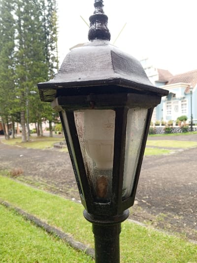 The image captures an outdoor lamppost in Weintiapa, Liberia, against a backdrop of foliage and architecture under an overcast sky. In the foreground, a black, weathered metal lantern-style lamppost stands vertically, centrally positioned. Its octagonal top cap culminates in a finial. The lantern body features multiple transparent glass panes, behind which a frosted white LED-style light bulb is visible. A small, reddish-brown insect or debris rests at the base of the bulb, inside the lantern. The glass panes and the lantern's metal frame exhibit visible dirt and grime.

To the immediate left of the lamppost, a strip of green grass borders a dark, textured paved pathway or road that curves gently into the mid-ground. Beyond the pathway, a line of tall, slender coniferous trees with dark green needles stands, partially obscuring a lighter-colored building further in the distance.

To the right of the lamppost, another section of green grass separates the foreground from a distinctive light blue building with a reddish-brown tiled roof. This building features white window frames and architectural details. Low-lying shrubs in dark planters are arranged in front of the building. The ground immediately surrounding the building appears to be paved or graveled. No people or animals are present in the scene.