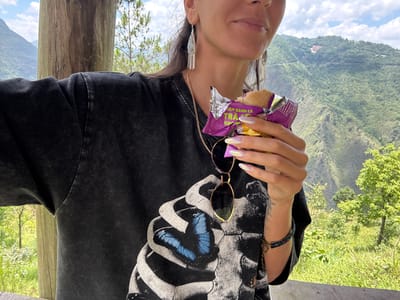 A woman is seen from a low angle holding a snack and sunglasses. She's wearing a dark shirt with a white skeletal-like design featuring a blue butterfly. She is also wearing earrings and a bracelet, and she has iridescent nails. The background features a green mountainous landscape and a blue sky with scattered clouds. This is in Lũng Cú Commune, Vietnam.