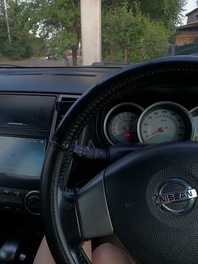The image shows the interior of a Nissan car, specifically the dashboard and steering wheel. Outside the car, through the windshield, a rural or suburban road is visible with trees and a building in the background. The car is likely stationary, as indicated by the unmoving gauges. The steering wheel is black leather with the Nissan logo in the center. To the left of the steering wheel is the car's infotainment system, displaying a music interface. Below that, there are climate control buttons and a gear shifter. The overall impression is that of a typical car interior on a slightly overcast day, possibly in a temperate climate. The specific mention of Belogorsk, Russia is not directly verifiable from the visual information alone, but if this context is provided, it suggests the car is located there.