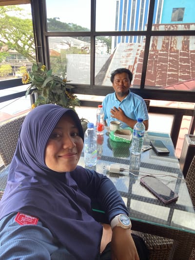 In an indoor cafe setting with a city view of Keraton, Indonesia, two individuals are seated at a glass-top table. The person in the foreground, a woman wearing a purple hijab and a blue long-sleeved shirt with an "EKSPEDISI 28 GUNUNG" patch, smiles at the camera. She wears a white wristwatch on her left arm. A mobile phone and a white charger are on the table in front of her. To her right, a bottle of Aqua water and a green tissue holder are visible.
In the background, a man in a light blue polo shirt is seated, with his hand on his chest. A cup and saucer, a bottle of water, and another mobile phone are placed on the table between them. The cafe has large windows offering a view of buildings with corrugated metal roofs and trees outside. The overall environment suggests a casual meeting or break.