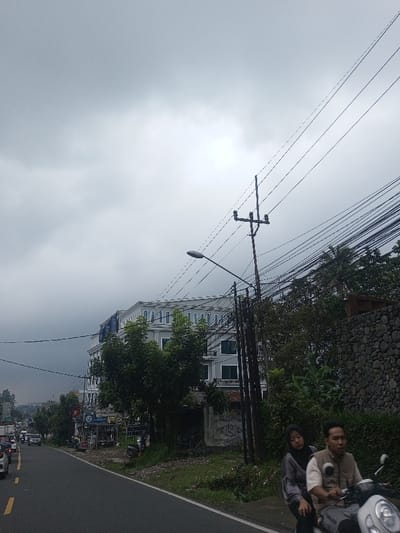 A street view beneath a cloudy sky features a white building partially obscured by trees. A utility pole stands tall, draped with numerous power lines. A stone wall is visible near foliage on the right. In the foreground, two people ride a scooter, moving from right to left. Traffic is seen on the left side of the road, which curves into the distance. A street lamp is present near the utility pole.