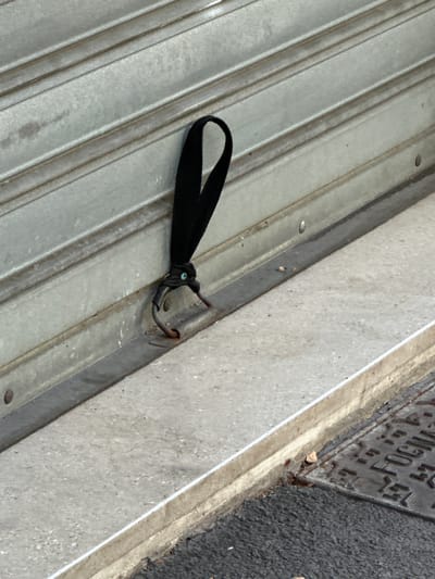 A black fabric loop is attached to a metallic, corrugated shutter. The loop is secured with a dark green string or cord, which is knotted around a dark metal hook or clasp that protrudes from the shutter. The shutter appears to be part of a shopfront or garage door. In the foreground, a concrete step or ledge runs horizontally across the image. To the right of the step, a textured dark surface, possibly asphalt, is visible, with a metal grate or manhole cover embedded in it, displaying partial lettering that reads "FOGNA". The overall scene suggests an outdoor urban environment in Trieste, Italy.