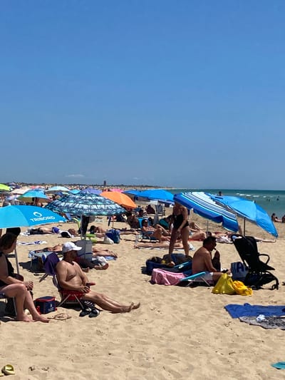 The image depicts a crowded beach scene at an unspecified location in Conil de la Frontera, Spain, on a sunny day. The sandy beach is dotted with numerous beach umbrellas of various colors and patterns, providing shade for a multitude of people lounging on towels and beach chairs. The ocean is visible in the background, with a few individuals in the water and a calm sea. The foreground shows people in swimwear, some sunbathing, others seated and relaxing. Objects such as coolers, bags, and a baby stroller are also present on the sand. The overall atmosphere suggests a typical summer day at a popular beach destination.