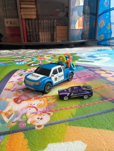A close-up, eye-level shot shows two toy cars on a colorful play mat. The play mat features cartoon characters like a teddy bear and a unicorn. In the background, a bookshelf is visible, filled with books.

The primary subject is a blue and white toy police truck, positioned in the foreground on the left. It has "POLICE HIGHWAY PATROL" written on its hood and a police badge on its door. On the back of the truck are several colorful, spiky toy gears.

To the right of the police truck and slightly in front of it is a small, purple toy car. This car has white racing stripes and "LIGHTNING" and "GPX" printed on its side.

The lighting appears to be natural, casting soft shadows. The overall scene is that of a child's play area. There are no people visible in the image. The location is not specified as Gornye Kljuchi, Russia.