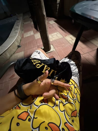 This image captures a low-light outdoor scene in Debelt, Bulgaria, depicting the lower torso, lap, and hands of an individual seated next to a swimming pool. The individual is wearing white undergarments with dark text, possibly "THUG," beneath yellow lounge pants or shorts adorned with a repeating cartoon duck pattern featuring black outlines and orange beaks. A black wristband or smartwatch is visible on the left wrist. The individual's hands are resting on a black velvet-like pouch with prominent gold embroidered lettering, partially visible as "VIP" or similar. A dark bottle, possibly containing liquid, is partially visible within or next to the pouch. A silver-colored ring with an intertwined design is on the ring finger of the left hand, and a gold-colored, patterned ring is on the index or middle finger of the right hand. The ground is paved with alternating reddish-brown and lighter-colored rectangular tiles. A dark, thick wooden post is present on the right, and the edge of a dark, textured outdoor table is visible in the upper right quadrant. A dark grey object, possibly an outdoor light fixture, is near the pool's edge. The scene is illuminated by unseen artificial light sources, casting shadows.