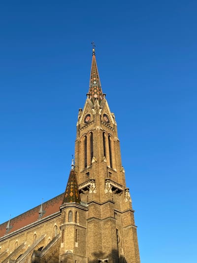 The image shows the upper portion of the Saint Teresa of Avila Cathedral in Subotica, Serbia, viewed from a low angle against a clear blue sky. The cathedral's architectural details are visible, including the spire, clock faces, and brickwork. The multi-colored tile roof is also prominent. There are no people visible in the frame.