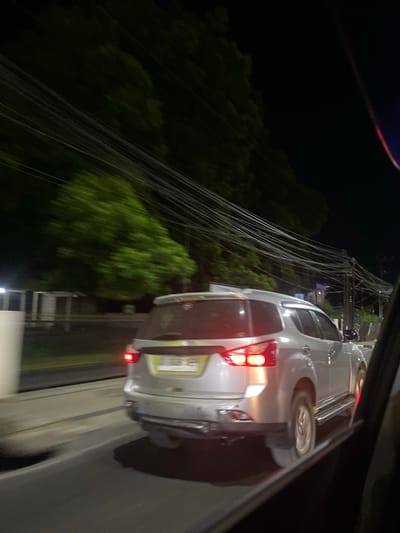 The image shows the rear of a silver SUV at night in Sungai Paring, Indonesia. The vehicle is parked on a paved road, with dense green trees on the left side of the frame. A tangle of electrical wires hangs precariously over the scene, obscuring some of the background and suggesting a somewhat cluttered or underdeveloped area. The car's taillights are illuminated, indicating it is either stopped or in motion. The lighting suggests it is nighttime, and the darkness of the sky and the illuminated car lights are prominent. There is no visible text in the image. The overall impression is a somewhat ordinary scene of a car at night, with the electrical wires adding a distinctive visual element.