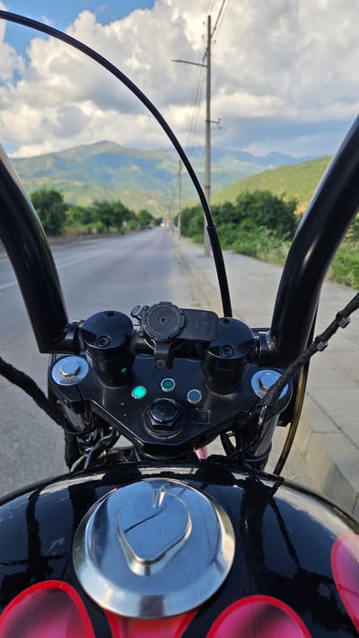 The image is taken from the perspective of a motorcycle rider on a paved road in Rila, Bulgaria. The handlebars of the motorcycle are visible in the foreground, with the dashboard and fuel tank also in view. The road stretches out ahead, lined with trees and leading towards rolling, green hills in the distance under a cloudy sky. A telephone pole stands on the right side of the road. The scene appears to be during daylight.