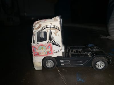 A white and black toy semi-truck cab and chassis is positioned on a dark, slightly reflective surface. The white cab of the truck shows significant wear, with brown, dirt-like residue covering the upper and front portions, and visible paint chipping. A large, black Mercedes-Benz three-pointed star logo is visible on the side of the cab, partially obscured by the dirt and wear. A rectangular sticker featuring the Sanrio character Cinnamoroll, a white dog with long ears, peeking from a pink-framed window with a small yellow chick on its head, is adhered to the lower front side of the cab. The truck's chassis is black, with molded side details. Two silver-colored wheels with black tires are visible: one beneath the cab and one at the rear of the chassis. The background is dark and out of focus, providing no discernible details. The specific location in the city of Karangkemiri, Indonesia, cannot be determined from the visual information presented in the image.