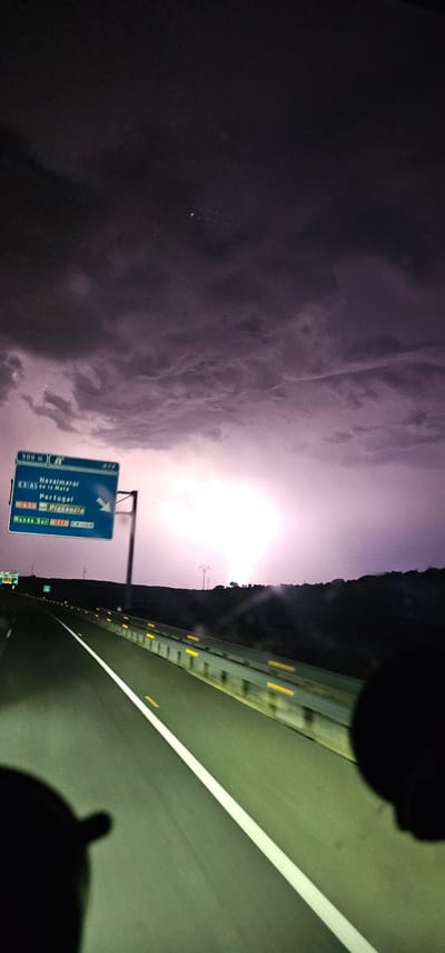 The image captures a highway scene at dusk or night, characterized by dramatic storm clouds and a bright lightning strike illuminating the sky. A road sign indicates directions to "Navalmoral" and "Portugal", with "Plasencia" also visible. The highway stretches forward, with lane markings and guardrails visible. The overall atmosphere is dark and stormy, with the lightning strike creating a powerful visual contrast against the dark sky.