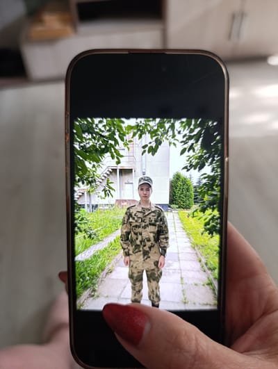 A person wearing a camouflage military uniform and a cap is standing on a paved path outdoors. The path is flanked by lush green grass and trees. In the background, there is a building with an external staircase and a tall, conical evergreen tree. The image is displayed on a smartphone, held by a hand with red nail polish. The overall scene appears to be a photograph taken in an outdoor, likely institutional setting, possibly a military facility, in Tugulym, Russia.