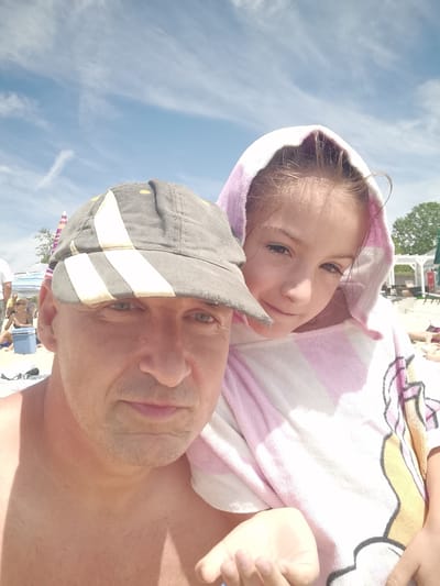 The image is a selfie taken outdoors on a sunny day, likely at a beach in Kranevo, Bulgaria. The foreground features two subjects: a man wearing a grey baseball cap and a young girl wrapped in a pink and white striped towel with a hood. The man is in the lower left corner, looking directly at the camera with a neutral expression. The girl is positioned behind and to the right of the man, with her face visible as she looks slightly off-camera with a gentle smile. Her hair appears damp, suggesting they may have been swimming.

In the background, the scene depicts a beach environment. There are beach umbrellas, lounge chairs, and other beachgoers, indicating a lively atmosphere. The sky is blue with scattered white clouds. The lighting suggests it is daytime, likely late morning or early afternoon. The overall mood appears relaxed and familial, as if capturing a moment of leisure during a vacation. No visible text is present in the image.