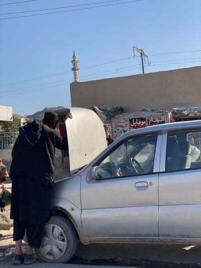 In کوکران, Afghanistan, a daytime scene depicts a roadside vehicle inspection or repair. A man, wearing a black tunic and a black cap, stands facing the open hood of a dirty silver compact car, holding the hood up with both hands. His back is mostly to the viewer. Partially visible inside the car, a second individual's hand and sleeve are seen reaching into the engine bay from the passenger side. A third person is partially obscured on the far left. The car's front driver's side wheel is visible, and the ground beneath it appears dusty. In the background, a light-colored minaret with a pointed spire rises above a beige wall. Along the middle ground, behind the car, are several makeshift market stalls or shops. One stall displays a sign in Arabic/Dari script reading "الهام موبایل" (Elham Mobile), indicating a mobile phone business. Overhead, multiple electrical lines traverse a clear blue sky, with a utility pole visible to the right. Distant mountains are faintly visible on the left horizon.