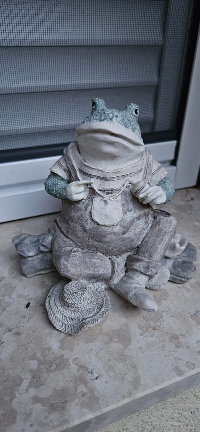 A static, full-shot, eye-level outdoor photograph shows a weathered, grey garden statue of a frog dressed in overalls and a shirt. The frog is seated with its legs crossed, holding small objects in each hand, and a miniature straw hat rests beside its left foot. The statue sits on a marble-like surface, possibly a patio or windowsill. Behind the statue is a white window frame and a grey, slatted screen. The lighting is bright and appears to be natural daylight, casting subtle shadows. The background is blurred, suggesting a shallow depth of field. The overall scene is still and devoid of any human presence. This scene is located in Hévíz, Hungary.