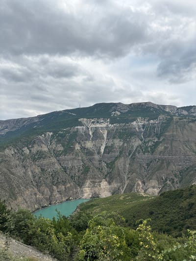 The image depicts a wide, scenic view of a canyon with a turquoise river flowing through it. The canyon walls are steep and rugged, with layers of rock and sparse vegetation. The upper slopes of the mountains are covered in a denser green forest. The sky is overcast with gray clouds, creating a subdued lighting condition. The foreground shows a hillside with a rocky path and lush green bushes and trees. A communication tower is visible on the highest peak in the background. The location is identified as Zubutli, Russia. There are no people or animals visible in the scene. The image captures a natural landscape characterized by its geological features and the presence of water and vegetation under a cloudy sky.