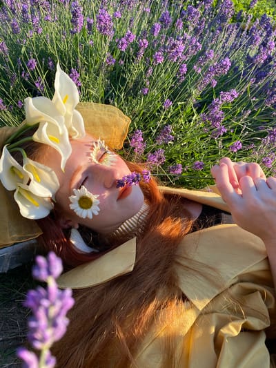 This media file depicts a close-up, overhead shot of a person lying in a field of lavender flowers. The person has reddish-brown hair and is adorned with flowers placed on their face, including a daisy over one eye and a cluster of lavender sprigs on the bridge of their nose. White calla lilies are also arranged around their head. The person is wearing a golden-colored garment that drapes around them. Their eyes are closed, and their expression appears peaceful or serene.

The scene is set outdoors, likely in a lavender field, given the abundance of purple flowers and green stems visible throughout the image. The lighting suggests it is daytime, possibly late afternoon or early morning due to the warm, golden hues cast on the fabric and hair. The presence of flowers and the serene pose evoke a sense of nature, tranquility, and perhaps artistic expression or a connection with nature. The image seems to capture a moment of quiet contemplation or rest amidst the natural beauty of the lavender field. No text is visible in the image.