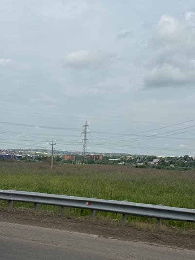 The image shows a landscape view under a cloudy sky, likely from the side of a road in Solontsy, Russia. In the foreground, a gray metal guardrail runs horizontally across the bottom of the frame, supported by concrete posts. Adjacent to the guardrail is a dirt shoulder and what appears to be asphalt road. Beyond the guardrail, a large field of tall green grass stretches into the midground. In the distance, a town or village is visible, characterized by numerous buildings with red-tiled roofs and some modern structures, including a tall brick building. Several power poles and high-voltage power lines crisscross the sky and extend across the landscape. One prominent, tall lattice steel power pylon stands in the mid-right of the image. The sky is overcast with scattered white and gray clouds, suggesting a generally diffused light. No people are present in the image.