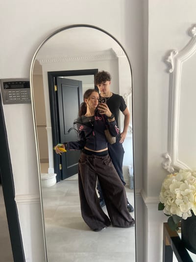 A reflection in a large, ornate mirror shows a young couple posing for a selfie. The man is standing behind the woman, both wearing dark clothing. The woman is holding a phone and a banana, appearing to take the photo. In the background of the reflection is a black door with gold trim, a white wall with decorative moldings, and what appears to be an entry intercom system. The mirror is set against a white wall and is flanked by an arrangement of white flowers on a table. The location is Welwyn Hatfield, United Kingdom.