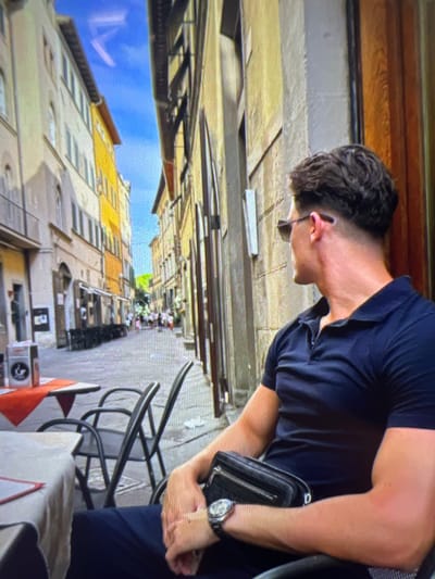 A man wearing a dark blue polo shirt and sunglasses is seated at an outdoor cafe table, facing away from the camera. He has a dark, textured bag slung across his waist and a watch on his left wrist. The cafe is situated on a cobblestone street lined with tall buildings painted in yellow and cream. In the background, several other patrons and cafe tables are visible, and the street leads to a bright sky. The scene appears to be in a European city, likely Italy, given the architecture and cafe setting. It is daytime, and the weather seems warm and sunny.