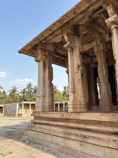 The image shows a perspective view of a structure in Kamalapura, India. The structure is characterized by pillars and stone construction, suggesting an architectural style typical of the region. The building materials are weathered, indicating age and exposure to the elements. The frame includes views of palm trees and other vegetation, suggesting a location in a tropical or subtropical area. The sky is visible through openings in the structure. There are no people visible.