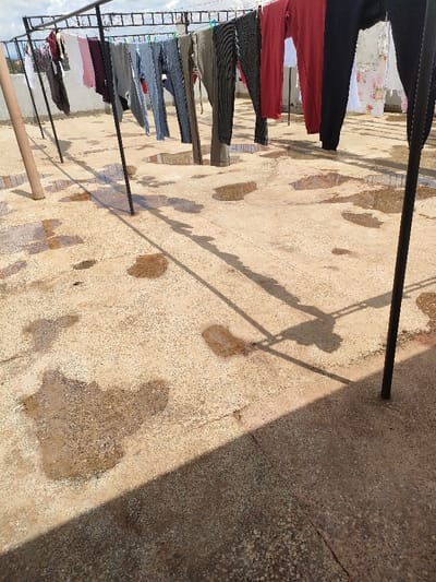 The scene depicts an outdoor concrete rooftop or elevated flat surface in Juja ward, Kenya, under bright sunlight. A metal drying rack, composed of dark vertical and horizontal bars, extends across the frame, supporting various articles of clothing suspended by clothespins. Garments include dark trousers/leggings (black, grey, olive green), maroon and white tops, and one white item with a floral pattern, all appearing to be in a state of drying. The concrete ground below is uneven and covered with numerous irregularly shaped puddles of water, suggesting recent rain or manual wetting. Distinct, elongated shadows of the drying rack and hanging clothes are cast across the wet surface, indicating a clear, sunny day. The background reveals a light blue sky with scattered white clouds and indistinct distant structures or terrain. No people or animals are present.