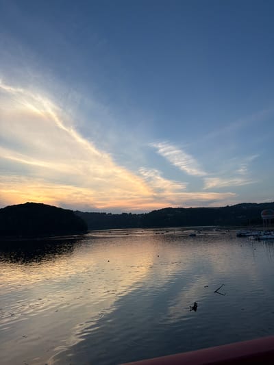 This image captures a serene sunset over the Rożnów Lake in Gródek nad Dunajcem, Poland. The sky is a gradient of deep blue at the top, transitioning to pale orange and yellow hues near the horizon, illuminated by wispy, elongated clouds. The calm lake reflects the colors of the sky, creating a shimmering, tranquil surface.

On the left side of the frame, a dark, silhouetted hill, densely covered in trees, juts into the water. The opposite shore is lined with more rolling, tree-covered hills, also in silhouette. Scattered across the lake are various boats, some anchored and others in motion, with a small cluster of them docked near the right bank. A colorful hot air balloon is visible on the far right, adding a touch of vibrancy to the scene.

The water's surface is slightly rippled, with reflections of the sky and distant landmasses. A few small objects, possibly debris or buoys, are visible in the water, and a distinct set of concentric ripples suggests something has recently disturbed the surface. The foreground shows the edge of a dark red railing, indicating the viewpoint is from a structure overlooking the lake. The overall atmosphere is peaceful and picturesque, highlighting the natural beauty of the location at dusk.