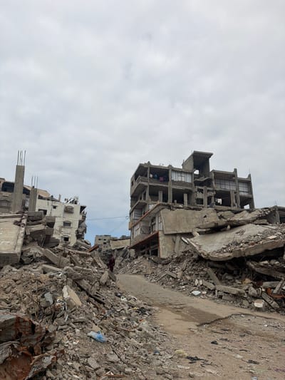 Rubble and collapsed buildings dominate the scene under a cloudy sky. Concrete debris is scattered across the foreground and background, indicating widespread destruction. The skeletal remains of structures rise from the debris, with exposed rooms and floors visible. The destruction suggests a war zone or a disaster area, conveying a sense of devastation.